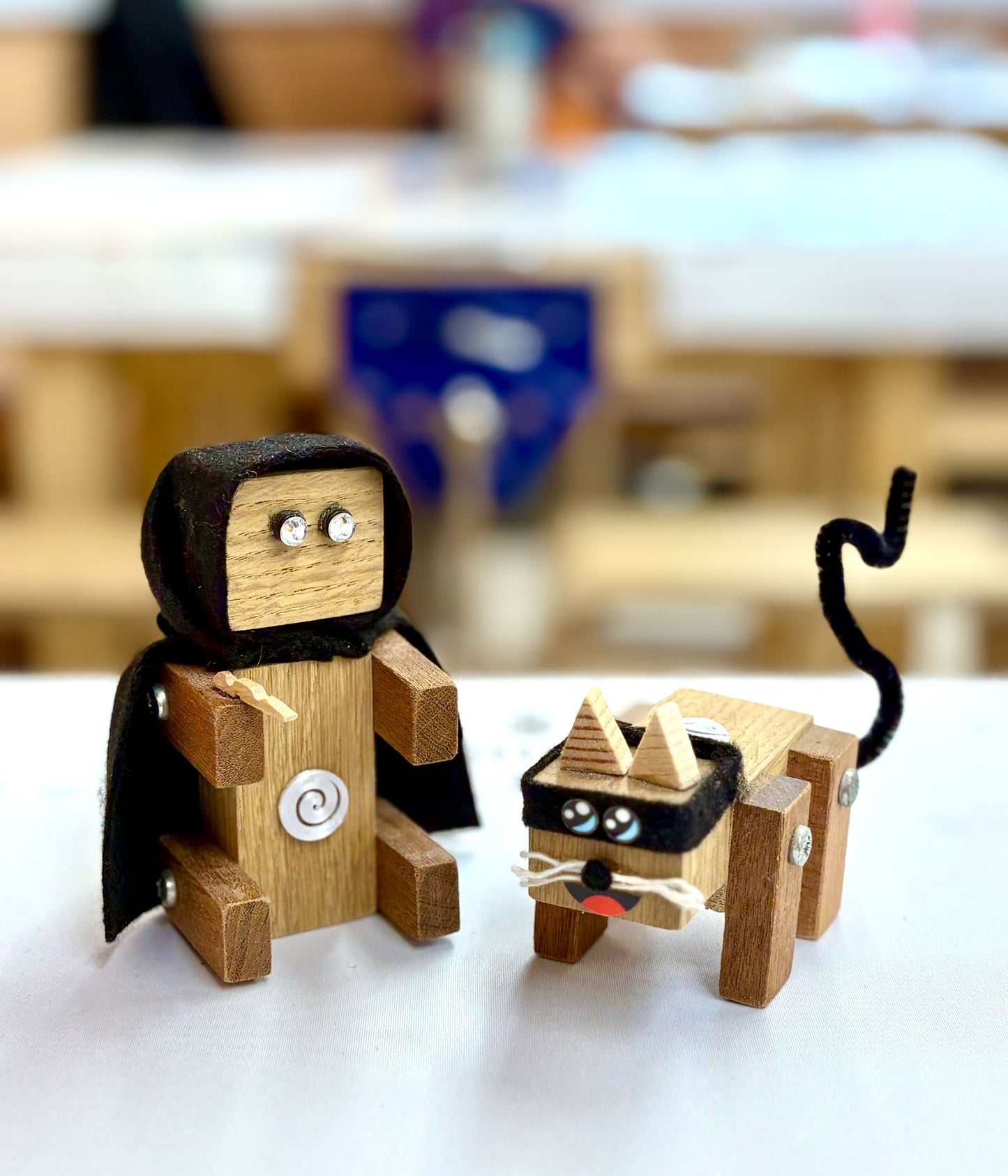 Two wooden super villain toys made at Ash and Co Workshops on a white surface with a blurred classroom background