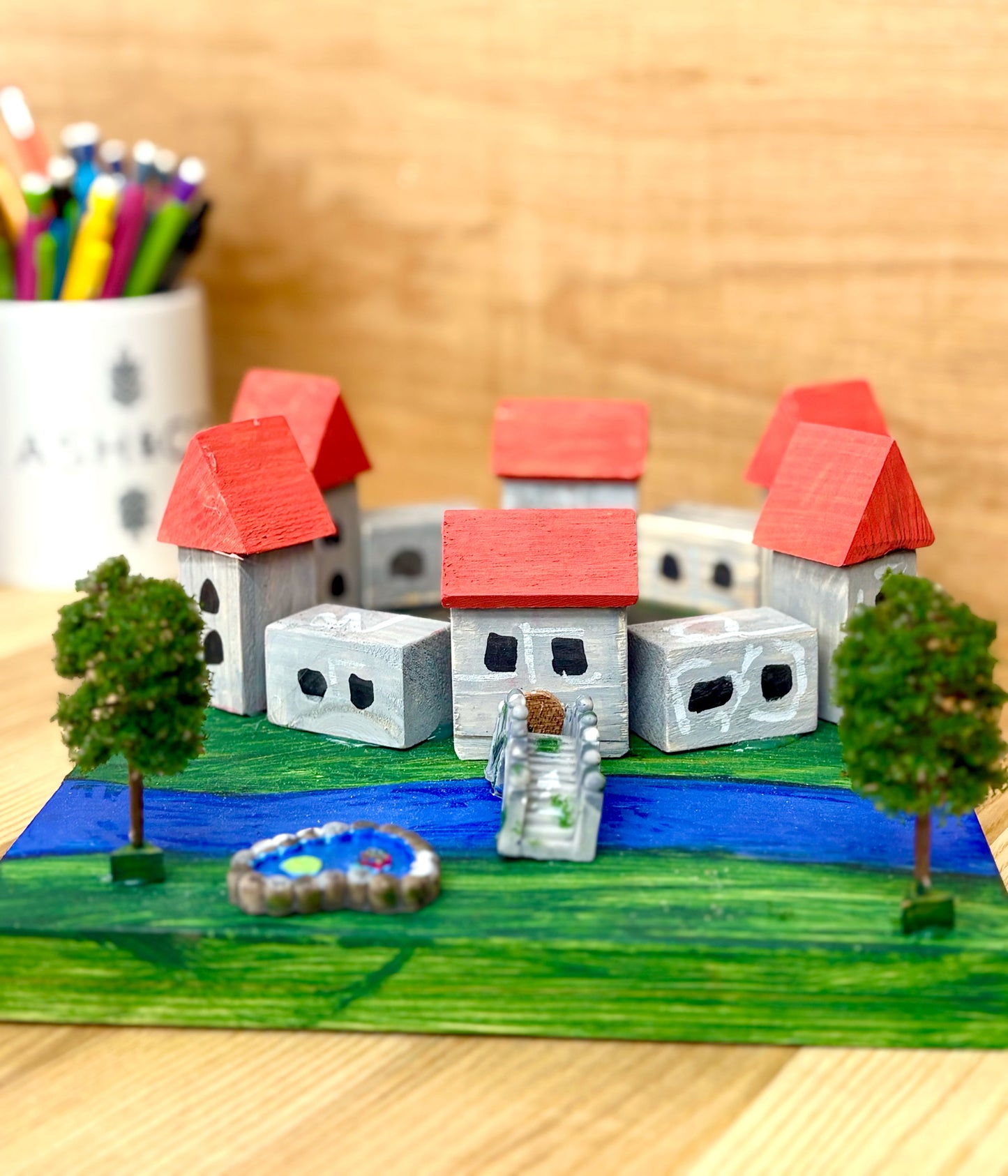 Model castle with houses, trees, and a pond on a wooden surface.