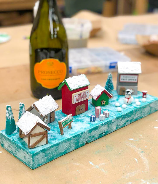 Miniature model of a snowy village scene on a table with a bottle of Prosecco in the background made at an Ash and Co Workshops Make n Sip Christmas event.