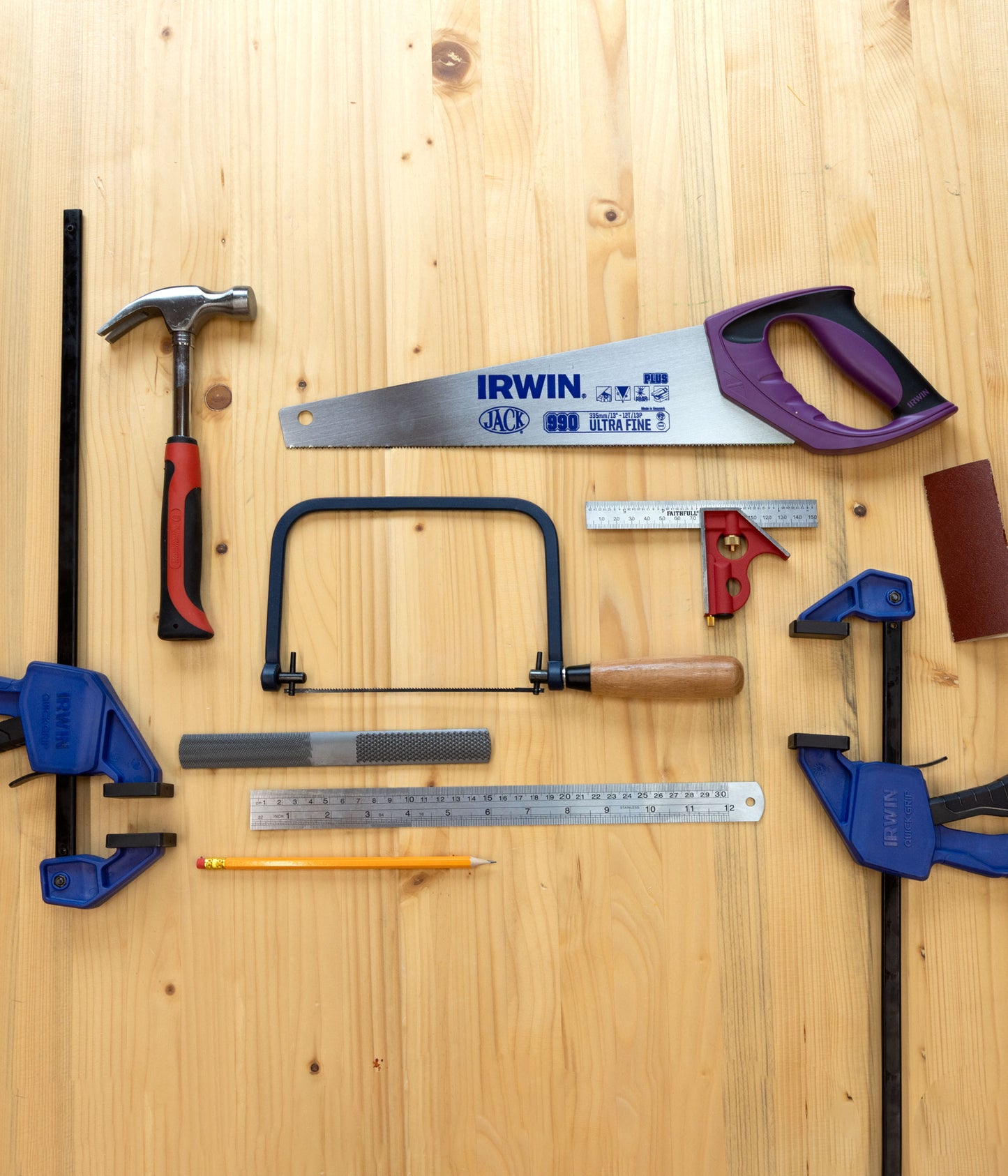 A flat-lay arrangement of various hand tools on a light wood background, including an IRWIN hand saw, a claw hammer, two blue clamps, a square, a ruler, a pencil, and other woodworking accessories.