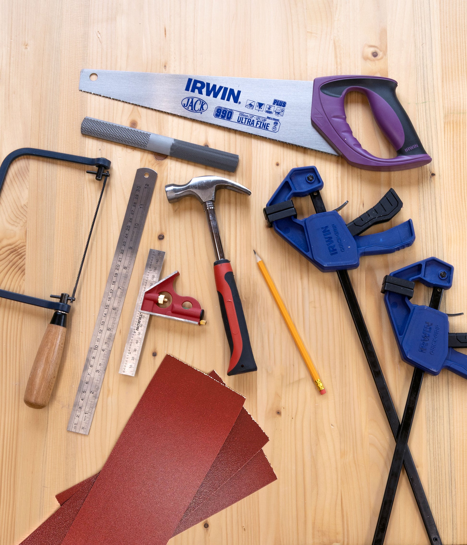 A flat-lay arrangement of various woodworking and carpentry tools on a light wood background, including an IRWIN hand saw, blue clamps, a hammer, a saw guide, a square, a pencil, and sheets of sandpaper.