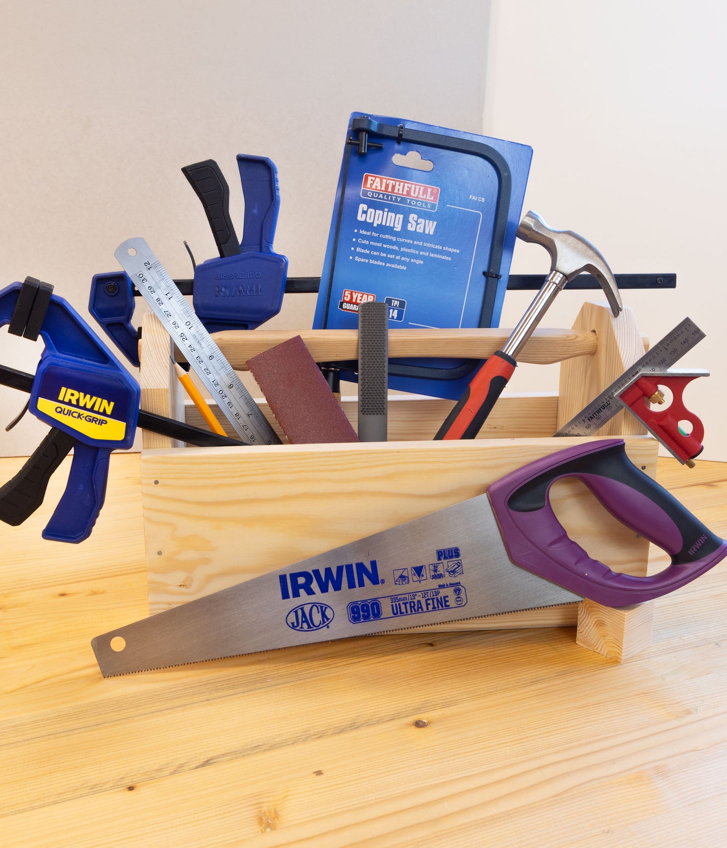 A wooden tool box filled with various hand tools, including an IRWIN jack saw in the foreground, a Coping Saw, clamps, rulers, and a hammer, set on a light wood surface against a plain light wall.