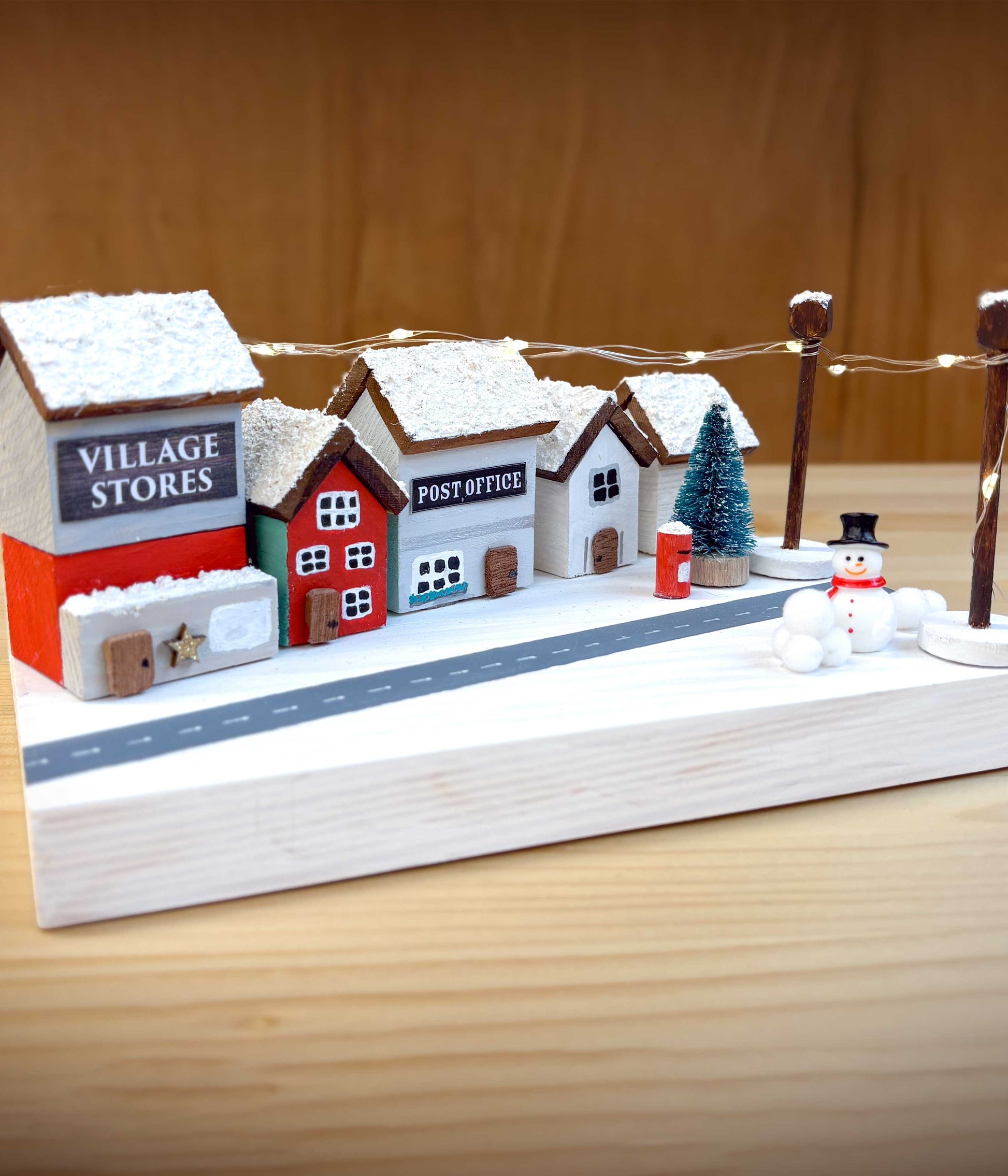 Wooden Christmas Village craft kit for kids - image shows completed model village scene with snow-covered roofs and a snowman on a wooden base.